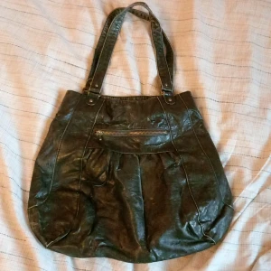 Svart axelväska i skinn med dragkedja - Stor svart axelväska i skinnimitation med coolt rutigt innerfoder. Väskan har dubbla axelband, en dragkedjeficka framtill och rymligt huvudfack med lite småfack också. Den är dock ganska sliten och på vissa ställen har skinnimitationen börjat gå bort (se sista bilden). 