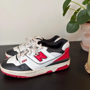 New Balance 550 sneakers vit/röd - Snygga New Balance 550 sneakers i vitt, svart och rött med klassisk retro vibe. Skorna har perforerade detaljer, röd N-logga på sidan och robust röd sula. Ovandelen är i skinn och textil, med snörning och låg profil. Perfekta för dig som gillar streetwear och sportig stil.