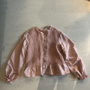 Ljusrosa blus med volang från Zara - Supersöt ljusrosa blus från Zara med smockad framsida, volangkant nedtill och vid halsen. Långa puffiga ärmar med resår och knappar framtill. Perfekt för dig som gillar romantiska detaljer och en feminin look. Har använt typ 3 gånger!!!