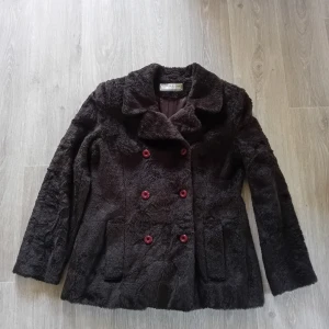 Mörkbrun pälsjacka med röda knappar - Säljer en mörkbrun kappa i fuskpäls I storlek 38. Kappan har 2 st fickor och knäpps med knappar som går åt det rödbruna hållet. Axel och nedåt ca 68cm lång och armhåla till armhåla ca 49cm 
