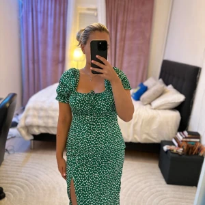 Grön prickig långklänning med puffärm - Supersöt grön långklänning med vita prickar, puffärmar och snygg slits framtill. Klänningen har knytning vid bysten och en figurnära passform som framhäver midjan. Perfekt för dig som gillar färg och mönster!