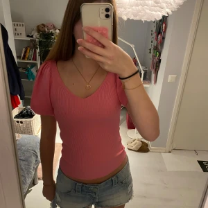 Rosa ribbad topp med puffärm och knyt - Supersöt rosa ribbad topp med v-ringning och puffärmar. Toppen har en snygg öppen rygg med knytband upptill som ger en extra detalj. Figurnära passform och kort ärm, perfekt till jeans eller kjol.