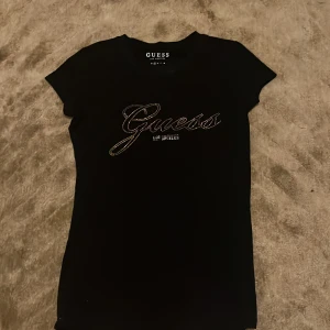 Svart t-shirt med strass från Guess - Svart t-shirt från Guess med korta ärmar och snyggt strassmönster i guld som bildar loggan på bröstet. Modellen är figurnära och har rund halsringning. Perfekt för dig som gillar stilrena plagg med en touch av bling.