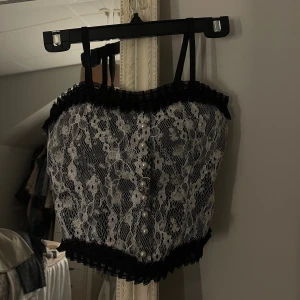 Svart spetskorsett med pärlknappar - Supersnygg svart korsett-topp med vit blommig spets och pärlknappar framtill. 