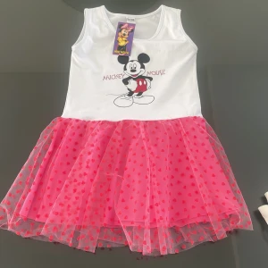 Mimmi Mouse klänning med hjärtan - Supersöt ärmlös klänning med vit överdel och tryck av Mickey Mouse. Kjolen är rosa med flera lager tyll och små röda hjärtan. Perfekt för dig som älskar Disney och vill sticka ut med en färgglad look.