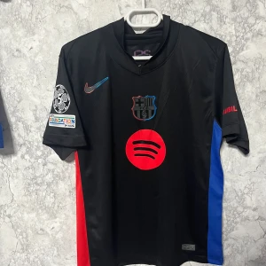 FC Barcelona Raphinha Nike fotbollströja - Snygg svart FC Barcelona fotbollströja från Nike med Raphinha och nummer 11 på ryggen. Tröjan har röda och blå detaljer på sidorna, klubbmärke och Spotify-logga framtill samt Champions League-märke på ärmen. Tillverkad i lätt Dri-FIT-material.