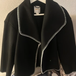 Svart omlottjacka från Gina Tricot - Svart omlottjacka från Gina Tricot med bred krage och vita dekorativa sömmar längs kanterna. Väldigt snygg och bekväm. Storlek 158/164 som motsvarar xs. Har dessutom knappar så man kan stänga den ifall man vill de😇