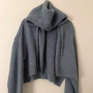 Blå stickad hoodie med dragkedja - Mysig blå stickad hoodie med huva och snörning. Tröjan är croppad och har en dragkedja framtill. 
