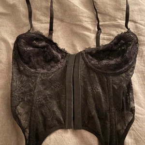 Svart spetskorsett med justerbara band - Säljer en svart korsett med vacker spets och blommigt mönster. Toppen har justerbara axelband och hakknäppning framtill. Perfekt för att skapa en edgy och trendig look. Sitter tight och formar kroppen snyggt.