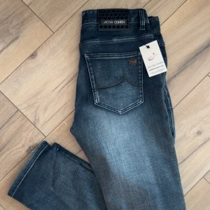 Jacob Cohen mörkgrå jeans straight fit 30 - Snygga mörkgrå jeans från Jacob Cohen med klassisk femficksdesign och diskret broderi på bakfickan. Jeansen har en rak passform och är tillverkade i mjukt denimtyg med lätt tvättad look. Perfekta för dig som gillar stilrena och exklusiva jeans. De är i storlek 30