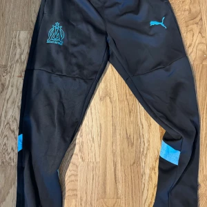 Svarta Puma träningsbyxor OM - Svarta träningsbyxor från Puma med Olympique de Marseille-logga i turkos på låret och matchande Puma-logga. Byxorna har smal passform, elastiskt material och turkosa detaljer vid vaderna. Perfekta för fotboll eller träning.