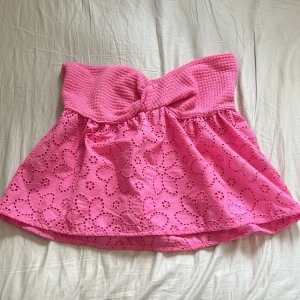 Rosa bandeau tubtopp - Supersöt rosa bandeau tubtopp med broderade blommor och hålmönster. Den är endast använd fåtal gånger. Toppen har en smockad överdel och en volangig, luftig nederdel. Perfekt för sommarens varma dagar och ger en riktigt fräsch vibe 💕
