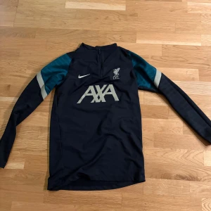 Liverpool tröja - Snygg långärmad Liverpool FC träningströja från Nike med halv dragkedja. Marinblå bas med turkosa och silverfärgade detaljer på axlarna. AXA-logga på bröstet och LFC-märke. Perfekt för träning eller att representera ditt lag. Materialet är lätt och andas.