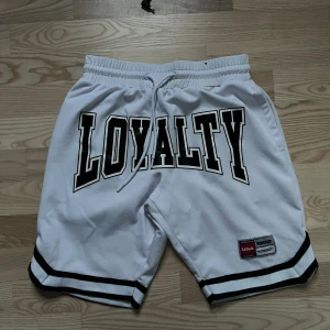 Vita basketshorts med LOYALTY-tryck - Snygga vita basketshorts med stort LOYALTY-tryck framtill och svarta ränder vid bensluten. De har snörning i midjan och en patch med USA Sports 1992 på benet. Perfekta för dig som gillar sportig streetstyle.