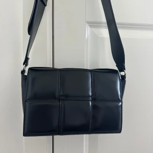 Svart quiltad axelväska från NAKD - Säljer en svart axelväska från NAKD med quiltad design och bred justerbar axelrem. Väskan är rektangulär och har flera fack inuti för smidig organisering. Tillverkad i glansigt syntetmaterial som ger en clean och modern look.