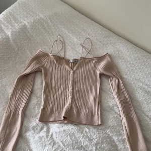 Beige ribbad offshoulder topp Dilvin - Trendig beige ribbad topp från Dilvin med offshoulder-design och smala axelband. Toppen har v-ringning, knappar framtill och långa ärmar med snygga cut-out-detaljer. Croppad passform som ger en modern vibe.