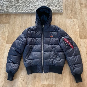 Mörkblå pufferjacka Alpha Industries - Snygg mörkblå pufferjacka från Alpha Industries med huva och klassisk röd tag på ärmen. Jackan har dragkedja framtill, ribbade muddar och två fickor med knappar. Perfekt för kalla dagar och riktigt street i stilen. Det är ett hål INUTI ena armen men inte så dunet åker ut och inte synligt utifrån. Annars helt felfri 