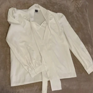 Vit blus med knyt från SHEIN - Säljer en vit blus från SHEIN med långa puffärmar och snygg knyt-detalj vid halsen. Blusen har en lätt glansig finish och klassisk krage. Perfekt för dig som gillar stilrena och trendiga plagg.