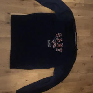 Mörkblå stickad tröja från GANT - Snygg mörkblå stickad tröja från GANT i storlek L. Tröjan har lång ärm och klassisk rund hals. På bröstet finns ett broderat GANT-tryck med amerikansk flagga och text. Perfekt för dig som gillar preppy stil och vill ha något bekvämt och stilrent.