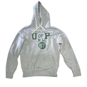 Ralph lauren hoodie - Snygg grå hoodie med tryck i grönt och vitt framtill där det står U OF P R.L. 67. Tröjan har huva med vit snörning och en stor magficka. Perfekt för chill dagar eller när du vill ha en sportig look.