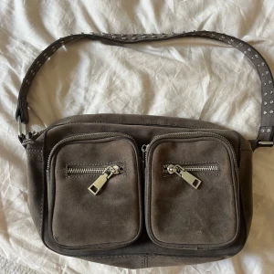 Brun grå mocka axelväska med nitar - Snygg brun axelväska i mocka med två stora framfickor och silvriga dragkedjor. Axelremmen är dekorerad med små nitar och det finns även en extra lång rem som kan bytas ut. Perfekt för dig som vill ha en cool och praktisk väska med plats för allt viktigt.