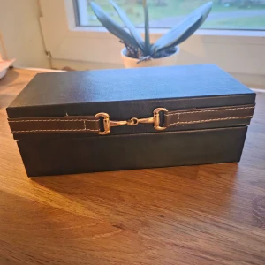 Svart smyckeskrin från Stjernsund kollektion  - Snyggt svart smyckeskrin från Stjernsund Kollektion med guldfärgad metallspänne och kontrastsömmar. Skrinet har en stilren, rektangulär form och är klädd i skinnimitation. Insidan är ljusbeige och rymlig. Mått 7 x 10 x 21 cm. Nyskick, aldrig använd. Endast upptagen ur förpackningen för att kunna fotografera den. 