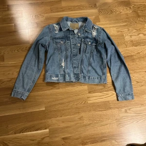 Blå jeansjacka med slitningar 157 - Säljer en blå jeansjacka från 157 med coola slitningar på axlar och framstycke. Jackan har klassisk krage, knappar framtill och två bröstfickor med lock. Perfekt för dig som gillar en avslappnad och trendig stil.