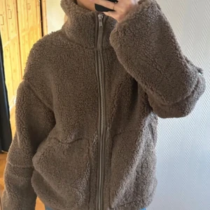 Brun teddyjacka med dragkedja - Mysig brun teddyjacka med hög krage och hel dragkedja framtill. Jackan har två stora fickor och en oversized passform som ger en chill vibe. Perfekt för kalla dagar när du vill vara både varm och snygg. Materialet är fluffigt och mjukt, vilket gör jackan extra skön att bära.