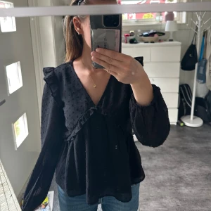 Svart blus med volang och prickar - Säljer en svart blus med v-ringning, volangdetaljer och diskret prickigt mönster. Blusen har långa ärmar med resår vid handlederna och en luftig, flowy passform. Perfekt att styla med jeans eller kjol för en trendig look.
