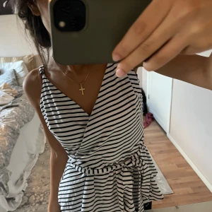 Randig omlott topp från Pagani - Supersöt omlott topp från Pagani i vitt med svarta ränder. Klänningen har smala axelband, v-ringning och knytskärp i midjan som ger en snygg siluett. Perfekt för varma dagar och enkel att styla med accessoarer.