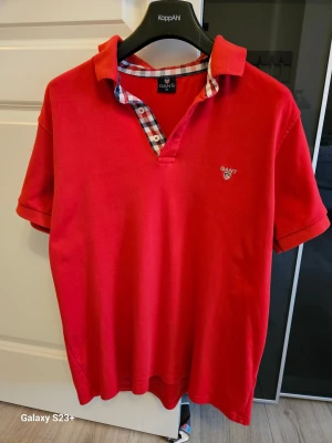Röd henley-skjorta från GANT XL - Röd henley-skjorta från GANT i bomull med klassisk krage och korta ärmar. Snygg rutigt mönstrad insida vid knäppningen och broderad GANT-logga på bröstet. Perfekt för dig som gillar stilren men ändå sportig look.