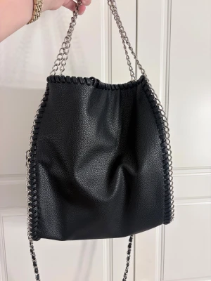 Svart axelväska med kedjedetaljer - Snygg svart axelväska från Gina Tricot i fejkskinn med silverfärgade kedjor längs kanterna och som axelrem. Väskan har ett rymligt huvudfack, innerficka med dragkedja och flera småfack. Perfekt för dig som gillar en edgy och trendig look.