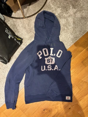 Blå hoodie från Polo Ralph Lauren - Snygg marinblå hoodie från Polo Ralph Lauren med stor vit print på bröstet där det står 'POLO 67 U.S.A.'. Tröjan har huva med snörning och en klassisk känguruficka framtill. Perfekt för dig som gillar sportig och avslappnad stil.