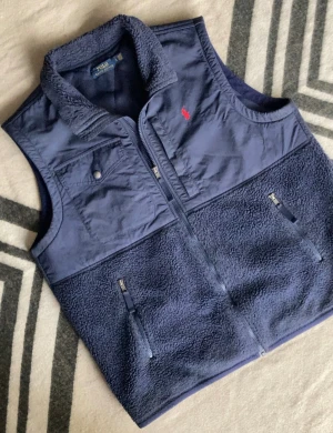 Mörkblå fleeceväst från Polo Ralph Lauren - Snygg mörkblå fleeceväst från Polo Ralph Lauren med mjuk teddytextur och detaljer i polyester. Västen har dragkedja framtill, två sidofickor med zip och en bröstficka med knapp. Klassisk röd Polo-logga på bröstet och hög krage för extra stil.