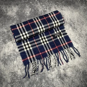Blårutig halsduk från Burberry - Snygg halsduk från Burberry i klassiskt rutig design med marinblå, vit, svart och röda detaljer. Gjord av 100% lammull, mjuk och varm med fransar i ändarna. Perfekt för att lyfta din vinterstil med lite brittisk vibe. Fråga gärna! Snabb leverans!