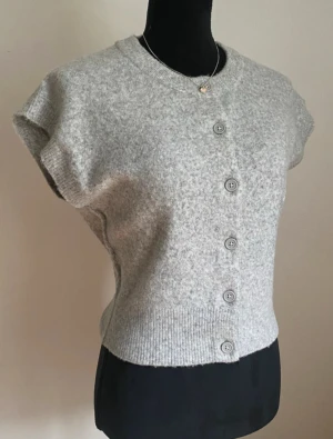 Grå kortärmad kofta från Nelly - Kortärmad grå kofta från Nelly med rund hals och fem knappar framtill. Koftan är stickad i mjukt material och har ribbade detaljer vid hals och nederkant. Perfekt för lager-på-lager och chill stil.