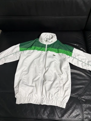 Grön och vit träningsdress från Lacoste - Retro träningsdress från Lacoste i grönt och vitt. Jackan har dragkedja, hög krage och två fickor, med gröna paneler över axlarna. Byxorna har resår i midjan och gröna samt vita detaljer längs sidorna. Klassisk krokodillogga på både jacka och byxa.