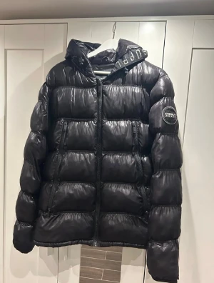 Svart pufferjacka Supply & Demand S - Svart pufferjacka från Supply & Demand med glansig finish, huva och dragkedja framtill. Jackan har två dragkedjefickor och logga på ärmen. Snygg detalj med text på huvan. Perfekt för kalla dagar och riktigt street.