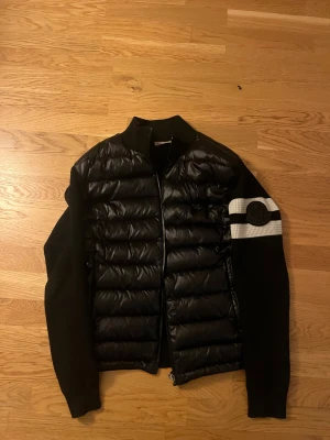 Svart Moncler pufferjacka med stickade ärmar - Svart pufferjacka från Moncler med stickade ärmar och quiltad framsida. Jackan har en vit och svart rand på ena ärmen med Moncler-logga, samt dragkedja framtill. Med funkande nfc tagg 