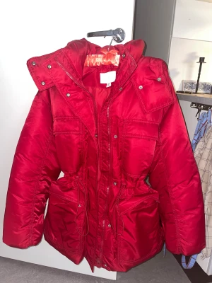Röd pufferjacka med huva och snörning - Säljer en röd pufferjacka med stor huva, snörning i midjan och flera praktiska fickor framtill. Jackan har dragkedja och tryckknappar, perfekt för kalla dagar. Materialet är glansigt och känns riktigt mjukt och skönt.