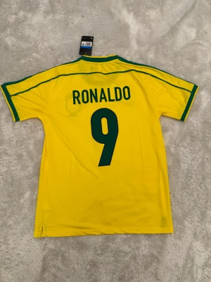 Brasilien Ronaldo 9 fotbollströja Nike M - Gul Brasilien fotbollströja med gröna detaljer, nummer 9 och Ronaldo på ryggen. Nike-logga och Brasilien-emblem med fem stjärnor på bröstet. Klassisk rund hals och korta ärmar. Tillverkad i lätt polyester med NikeFit-teknologi för skön känsla.