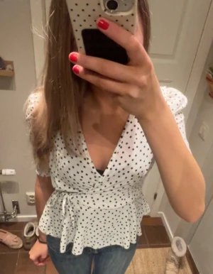 Prickig omlottblus från Boohoo - Säljer en vit omlottblus från Boohoo med svarta prickar, v-ringad hals och korta puffärmar. Blusen har knyt i midjan och volang nedtill för en extra söt look. Perfekt till jeans eller kjol för en trendig stil.