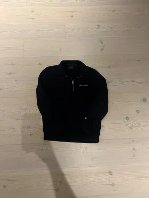 Mörkblå Half Zip Lyle&Scott - Mörkblå tröja från Lyle & Scott. Tröjan har en stilren design med broderad text på bröstet och är gjord i mjuk bomull. Perfekt för dig som gillar klassisk streetwear med en clean look. Storlek M