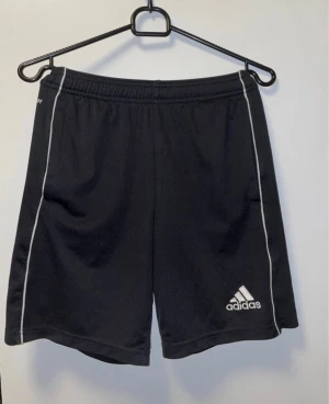 Svarta träningsshorts från Adidas - Svarta sportiga shorts från Adidas med vit logga och tunna vita linjer längs sidorna. De har elastisk midja och är gjorda i ett lätt polyestermaterial som andas. Perfekta för gymmet eller fotbollsplanen, klassisk stil med loose passform.