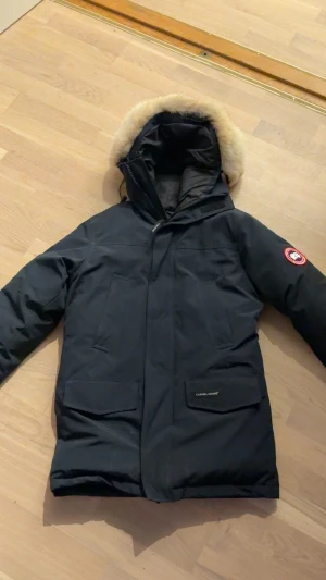 Svart Canada Goose parkas med päls - Svart parkas från Canada Goose med ikonisk patch på ärmen och stor huva med ljus päls. Jackan har stora fickor framtill, dragkedja och vindslå. Perfekt för kalla dagar med sin dunfyllning och klassiska look.