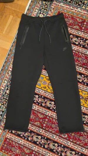 Svarta Nike mjukisbyxor med snörning - Svarta mjukisbyxor från Nike med dragsko i midjan och dragkedjeförsedda fickor. Byxorna har en rak passform och är tillverkade i ett mjukt syntetmaterial. Diskret Nike-logga på ena benet och justerbara benslut med snörning för extra komfort.