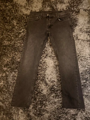 Svarta skinny jeans från Nudie Jeans - Säljer ett par svarta skinny jeans från Nudie Jeans med klassisk femficksdesign och snygga detaljer på bakfickorna. Jeansen har dragkedja och knapp framtill, smal passform och är tillverkade i mjukt jeanstyg. Perfekt för dig som gillar stilrena och tidlösa jeans. Jeansens är knappt använda (två gånger) Storlek: W31 L32