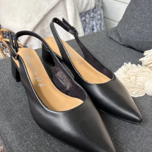 Svarta slingback pumps från Tamaris - Säljer ett par eleganta svarta pumps från Tamaris med slingback och låg klack. Skorna har spetsig tå och är helt oanvända. Perfekta för dig som gillar stilrena och klassiska skor.