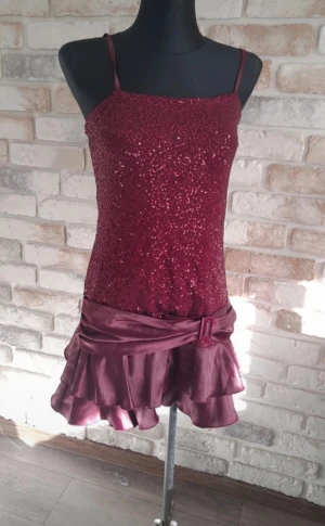 Vinröd glittrig kortklänning  - Vinröd kortklänning med smala axelband och glittrig paljettöverdel. Kjolen har dubbla volanger i satin och ett dekorativt bälte i midjan. Perfekt till jul. 