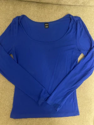 Kornblå långärmad topp från SHEIN - Säljer en kornblå långärmad topp från SHEIN med rundad, djup urringning. Modellen är slim och sitter snyggt figurnära. Materialet är mjukt och stretchigt, troligen bomull med inslag av syntet. Perfekt att styla med jeans eller kjol för en clean look.